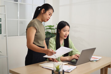 Portrait of Asian woman standing holding documents while discussing business ideas and plans with coworker sitting in front of laptop.