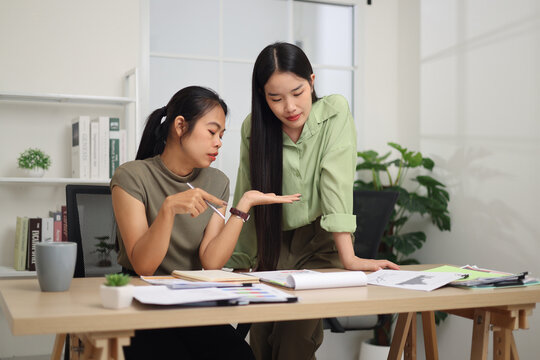 Portrait of Two female colleagues are working together in an office. A young woman is standing, talking and exchanging ideas with another colleague who is sitting.
