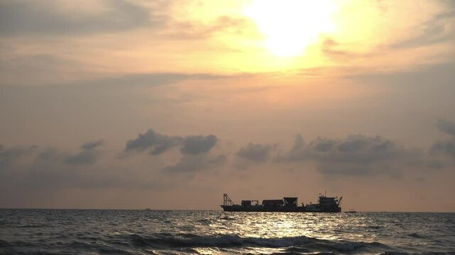 Silhouette vdo clip of a large ship floating on the ocean during a dramatic sunset. The sky glows in warm orange and yellow hues, reflecting softly on the rippling waves. A peaceful maritime scene