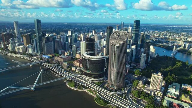 An aerial view captures Brisbane's city center with its winding river, bustling highways, towering skyscrapers, and iconic pedestrian bridges.