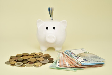 White piggy bank with banknote sticking out and stacks of coins and different value euro banknotes on the pale yellow background. Saving money and financial concept. Copy space. Selective focus.