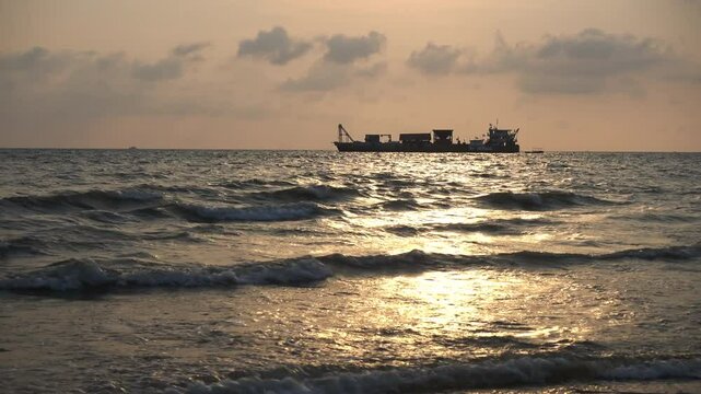 Silhouette vdo clip of a large ship floating on the ocean during a dramatic sunset. The sky glows in warm orange and yellow hues, reflecting softly on the rippling waves. A peaceful maritime scene