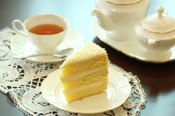 
A light and airy slice of white cake with creamy frosting and delicate crumbs is presented on a white plate with a doily, accompanied by a cup of tea and a classic white teapot