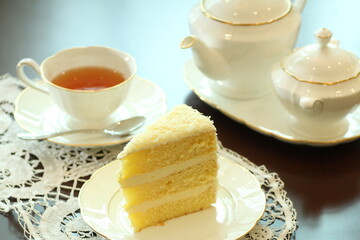 
A light and airy slice of white cake with creamy frosting and delicate crumbs is presented on a white plate with a doily, accompanied by a cup of tea and a classic white teapot