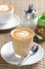 A steaming cup of latte art with a heart design sits on a wooden board next to a slice of cake and a vase of yellow flowers.