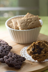 A rustic wooden board holds an assortment of delicious homemade cookies, including chocolate cut-out cookies, classic chocolate chip cookies on a doily, and plain square biscuits in a white fluted bow
