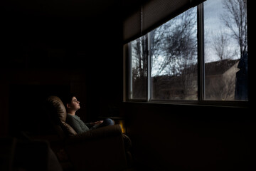 Tween boy sits in dark room in front of large window watching the rain