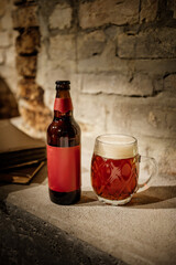 A brown glass beer bottle with a red label stands beside a full frothy mug of amber beer on a rough stone surface against a textured brick wall