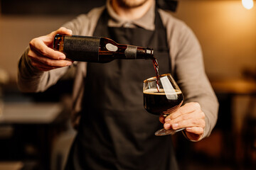 A bartender in an apron pours dark beer from a brown bottle into a tulip glass under warm indoor lighting. No visible logos or branding