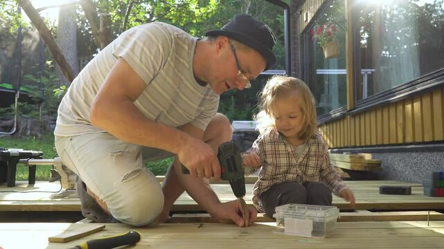 Father and his toddler work together on building a wooden terrace at home. The man uses a drill while the child observes and learns. Meaningful family moments. Give me a high five