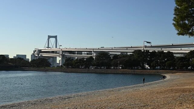 Calm and serene scene on the waters of Tokyo Bay with a alking person at Odaiba Beach with view on the Rainbow Bridge outdoor during sunset in winter in Tokyo city in Japan.