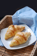  two golden-brown croissants on a white plate, resting on a wicker tray with a blue and white striped cloth