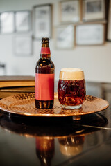 A frothy amber beer in a patterned glass sits beside a matching bottle with a red label on a hammered copper tray, with books and frames in background