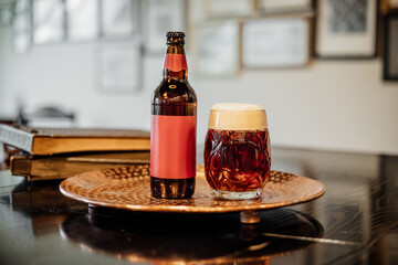A frothy amber beer in a patterned glass sits beside a matching bottle with a red label on a hammered copper tray, with books and frames in background