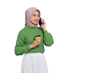 Portrait of smiling beautiful Asian woman in green sweater and hijab holding coffee cup while talking on mobile phone isolated on transparent background