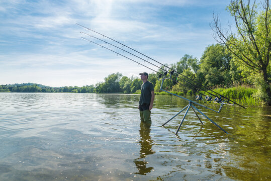 Fishing adventures, carp fishing. Man fishing on a lake in a beautiful sunny day. Fisherman standing, in the water with high boots, next to fishing equipment