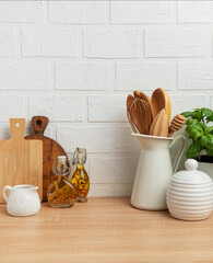 Kitchen utensils, basil plant and condiments resting on wooden countertop in front of white brick wall