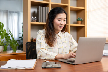 Young Asian woman wearing checkered shirt standing by wooden bookshelf arranging documents with smile, working from home in bright modern interior, productivity and organization concept.