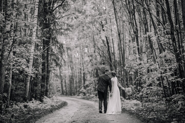 A romantic black and white photo of a newlywed couple kissing on a forest path, holding hands as they walk away into the woods..