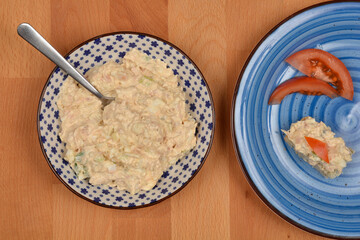 A small canape with tuna, cream cheese, egg, caper, onion, and mustard spread and tomato sits on a blue plate next to a bowl of the spread with a spoon