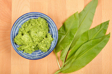 A blue and white bowl of vibrant green wild garlic (Allium ursinum) butter sits beside fresh wild garlic leaves on a wooden surface, captured from above