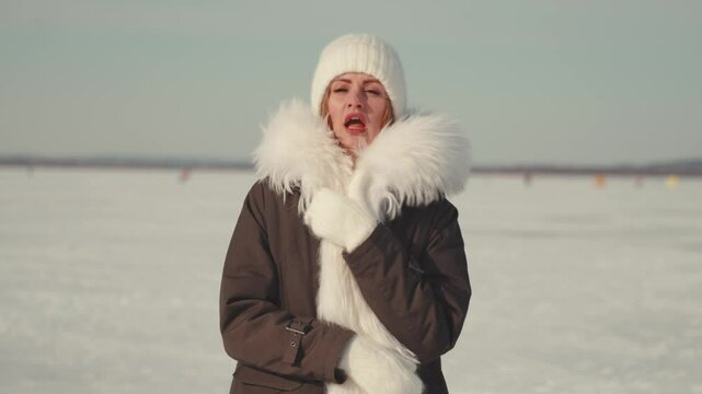 Frozen lake landscape provides the backdrop for a young woman experiencing extreme cold, bundled in winter clothing, visibly shivering and trying to warm herself