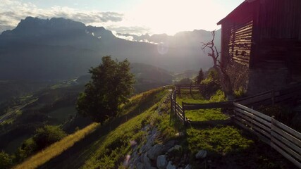Vertical drone ascent during a magical backlit sunset in the mountains of Tyrol, Austria. A sunlit mountain hut in golden hour contrasts with a valley road below. - Powered by Adobe