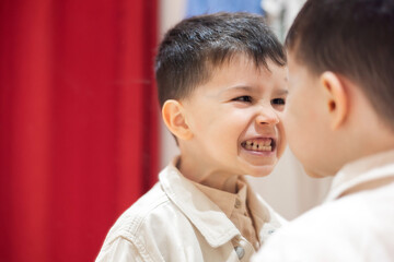 kid in fitting, dressing room trying on clothes. boy with stack of wears on hands sitting on chair. cute adorable child making funny faces in mirror.smiling baby in shopping center, mall.