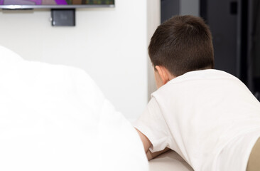 kid with remote in hand lying on bed watching tv. curios focused child boy wrapped in blanket. white curtains in background.top view. child with fist under chin.