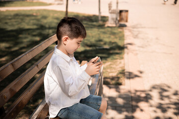 Fototapeta premium preschooler kid using smartphone sitting in park on bench.boy playing on mobile,taking selfie or registering a video.little blogger talking with his followers.child learning from tree.phone addiction