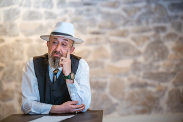 Elderly man in a white hat and vest gesturing expressively while sitting at a table, portraying a...