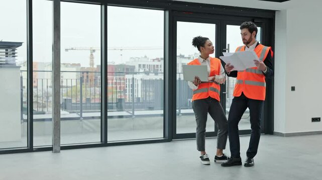 Two professional architects wearing safety vests are checking blueprints and discussing a project inside an empty office building.