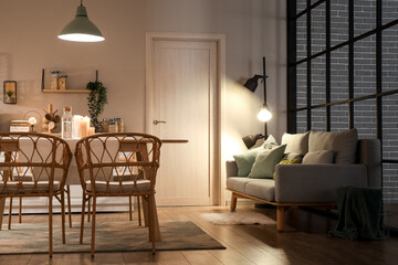 Interior of modern kitchen with glowing lamps, white counters, sofa and dining table at evening