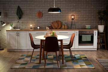 Interior of kitchen with glowing lamps, white counters, oven and dining table at evening