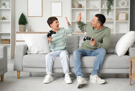 Cute little boy and his father with game pads giving each other high-five on sofa at home