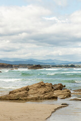 Coastal view with rocky outcrops and turquoise waves rolling onto a sandy beach, with mountains visible in the distance