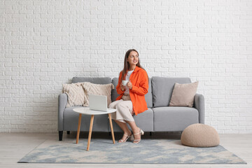 Obraz premium Young pretty woman with cup of coffee sitting on grey sofa at home