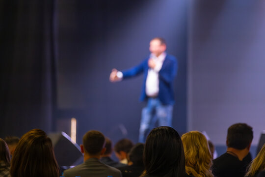 Presenter speaking on stage with blurry focus, addressing audience in a conference setting.