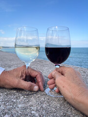 Two hands holding glasses of red and white wine with sea ocean view clouds reflected in wine  warm summer spring sunshine blue sky