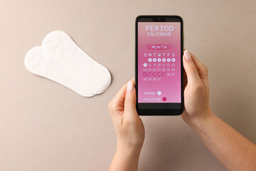 Menstruation. Woman holding smartphone with period calendar application on beige background, above...