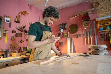 Smiling relaxed carpenter pauses work, comfortably checking smartphone news and unwinding before next handcrafted furniture commission, customers. Calm male relaxing in artisan workspace alone