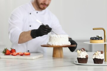 Confectioner decorating delicious cake at white marble table in kitchen, focus on different desserts