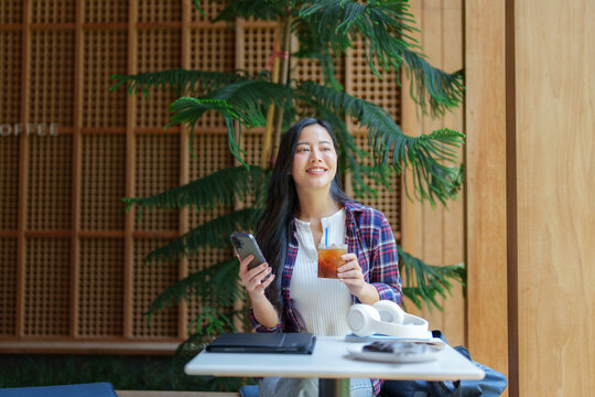 Young asian woman holding a smartphone and an iced coffee while working remotely at a table in a modern coffee shop, enjoying the pleasant atmosphere and taking a break - Powered by Adobe