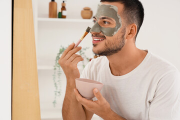Handsome young happy man applying cosmetic clay mask on face near mirror in bathroom