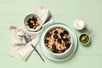 Bowl of tasty oatmeal with prunes, hazelnuts, honey and jug of milk on green wooden background