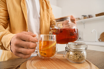 Young man pouring tea into glass cup with lemon on table in kitchen, closeup