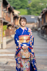 A teenage Japanese girl in a blue kimono standing in a traditional townscape of a regional city in Japan on a rainy summer day.