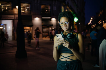 Happy woman checking phone at night in city center, using entertainment or gaming app, headphones...