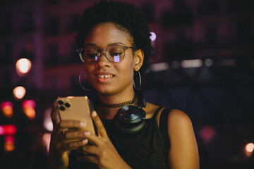 Urban Black woman looking at smartphone in nighttime city scene, screen light reflecting on face, wearing choker and over-ear headphones, possibly chatting or exploring online media