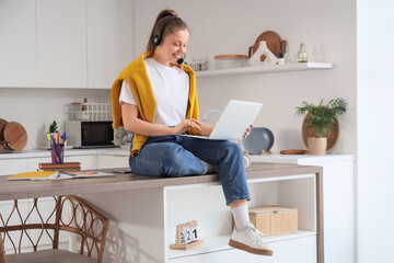 Teenage girl with headset and laptop working online in kitchen